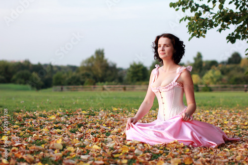 Fine emotional brunette sitting on autumn leaves and grass. Copy space. Yonge girl in vintage pink pastel with live expressions on beautiful country and old background.