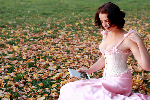 Fine emotional brunette with tablet pad sitting on the grass in autumn. Copy space. Yonge girl in vintage pink pastel with live expressions on beautiful country and old background.