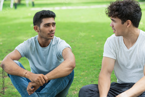 Serious Indian guy sharing problem with friend in summer park. Handsome young men sitting on grass and talking. Chatting concept