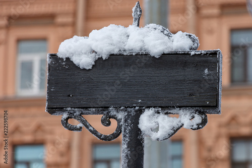 Vintage, black nameplate made of wood and wrought iron. The sign is covered with snow. On the background of an old light brown house. There is a place under copy space