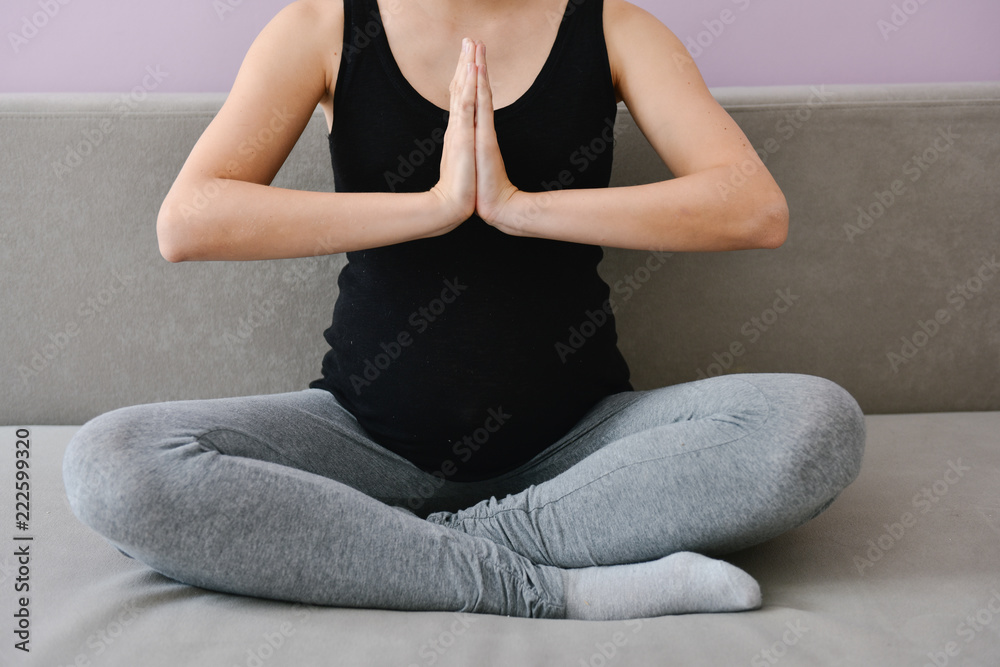 Cropped portrait of young pregnant woman meditating in lotus pose on sofa at home.