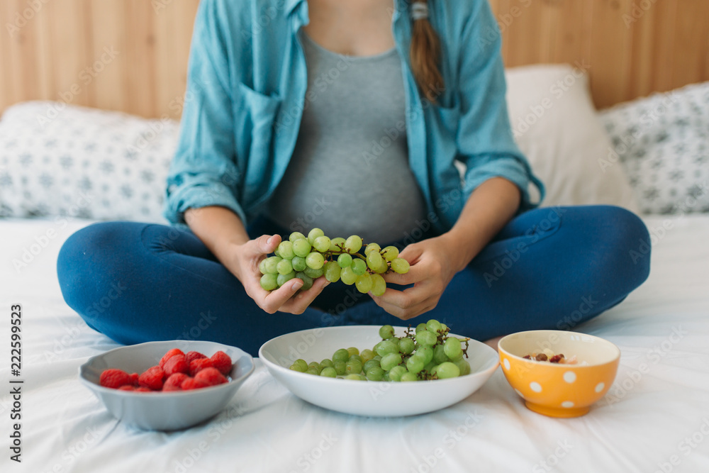 Cropped photo of young pregnant woman eating grapes, nuts and strawberries in bed Cropped photo of young pregnant woman eating grapes, nuts and strawberries in bed