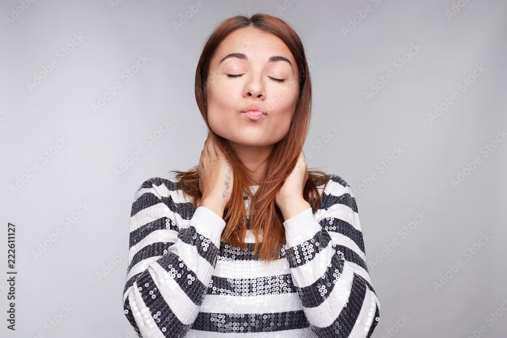 Fashionable young good looking female with dark ginger hair keeps lips round, looks coquette, keeps hands on neck, sends kiss at camera, poses against gray background, having fun and flirting look. Fashionable young good looking female with dark ginger hair keeps lips round, looks coquette, keeps hands on neck, sends kiss at camera, poses against gray background, having fun and flirting look.