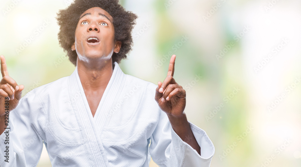Afro american man wearing karate kimono over isolated background amazed and surprised looking up and pointing with fingers and raised arms. Afro american man wearing karate kimono over isolated background amazed and surprised looking up and pointing with fingers and raised arms.