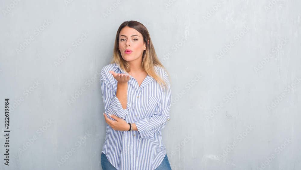 Young adult woman over grey grunge wall wearing fashion business outfit looking at the camera blowing a kiss with hand on air being lovely and sexy. Love expression. Young adult woman over grey grunge wall wearing fashion business outfit looking at the camera blowing a kiss with hand on air being lovely and sexy. Love expression.