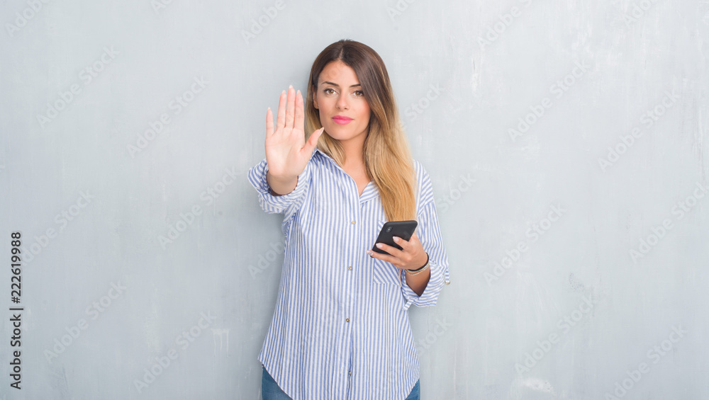 Young adult woman over grey grunge wall looking at smartphone texting a message with open hand doing stop sign with serious and confident expression, defense gesture Young adult woman over grey grunge wall looking at smartphone texting a message with open hand doing stop sign with serious and confident expression, defense gesture