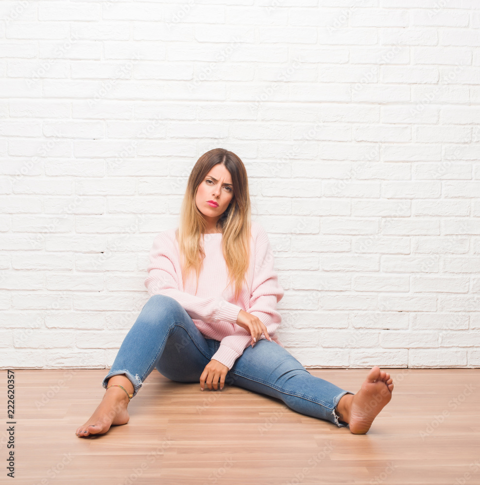 Young adult woman sitting on the floor over white brick wall skeptic and nervous, disapproving expression on face with crossed arms. Negative person.