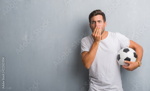 Handsome young man over grey grunge wall holding soccer football ball cover mouth with hand shocked with shame for mistake, expression of fear, scared in silence, secret concept