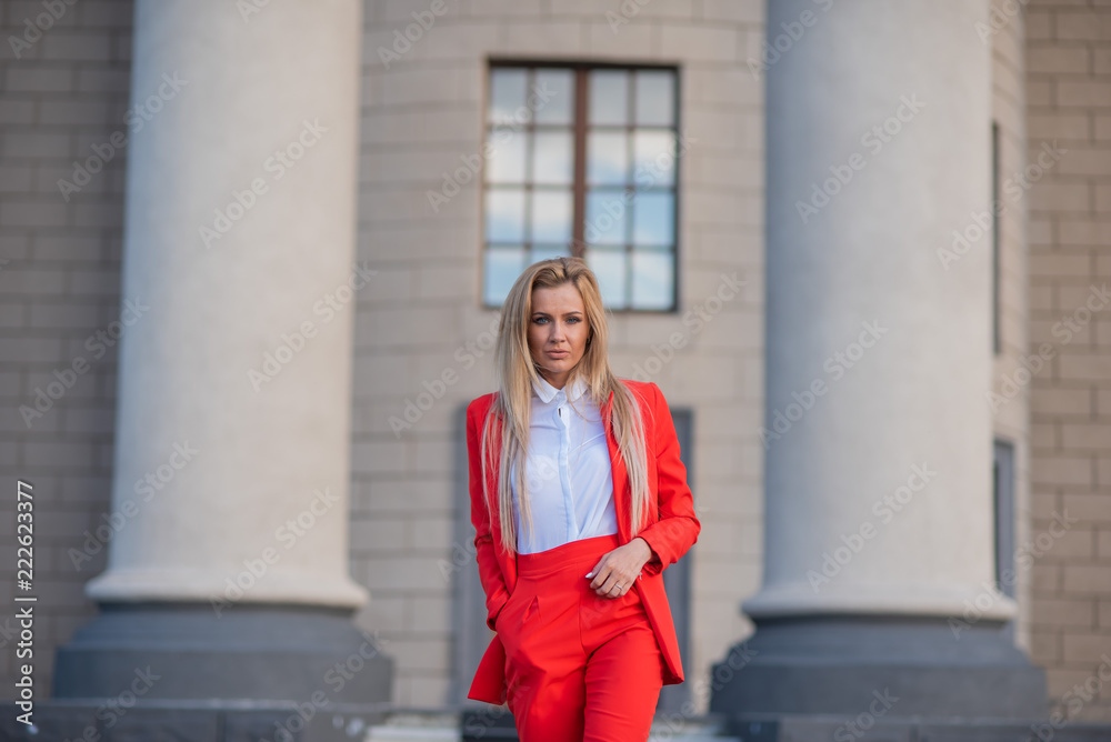 Beautiful blonde in a red jacket and pants on a building background with columns. A spectacular young woman in a trouser suit stands outside Beautiful blonde in a red jacket and pants on a building background with columns. A spectacular young woman in a trouser suit stands outside