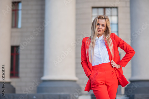 Beautiful blonde in a red jacket and pants on a building background with columns. A spectacular young woman in a trouser suit stands outside © Михаил Решетников