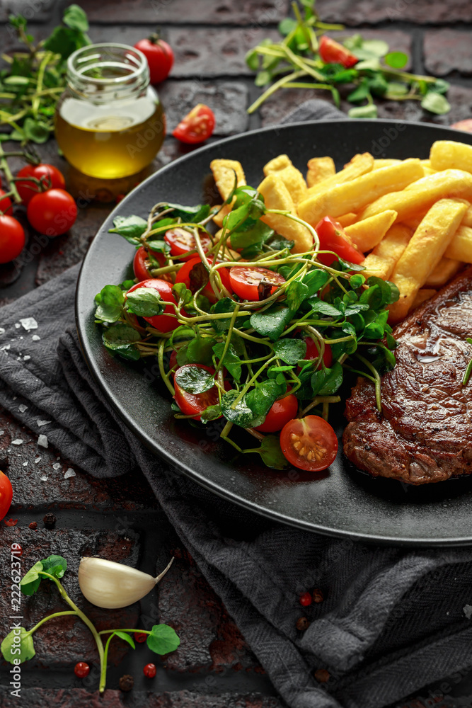 Grilled sirloin steak with potato fries and vegetables, tomato salad in a black plate. rustic table Grilled sirloin steak with potato fries and vegetables, tomato salad in a black plate. rustic table