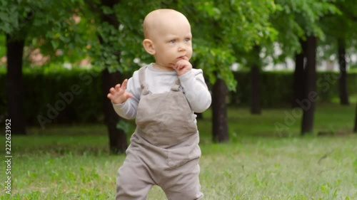 Tracking of fussy baby boy wearing overalls toddling around and taking his first steps on green grass in park