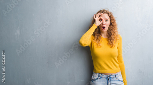 Young redhead woman over grey grunge wall doing ok gesture shocked with surprised face, eye looking through fingers. Unbelieving expression.