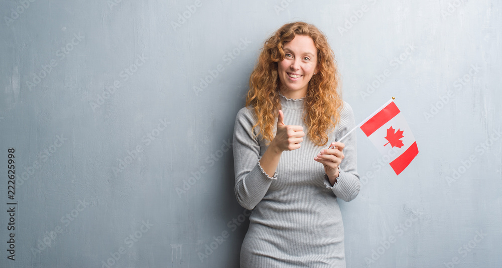 Young redhead woman over grey grunge wall holding flag of Canada happy with big smile doing ok sign, thumb up with fingers, excellent sign Young redhead woman over grey grunge wall holding flag of Canada happy with big smile doing ok sign, thumb up with fingers, excellent sign