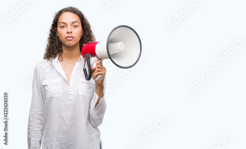 Young hispanic woman holding megaphone with a confident expression on smart face thinking serious