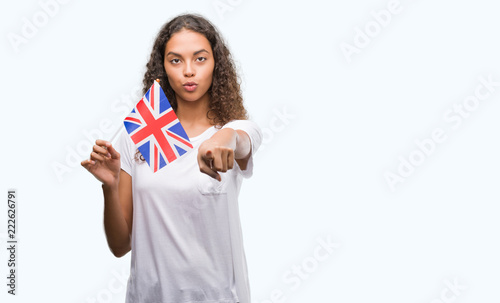 Young hispanic woman holding flag of United Kingdom pointing with finger to the camera and to you, hand sign, positive and confident gesture from the front