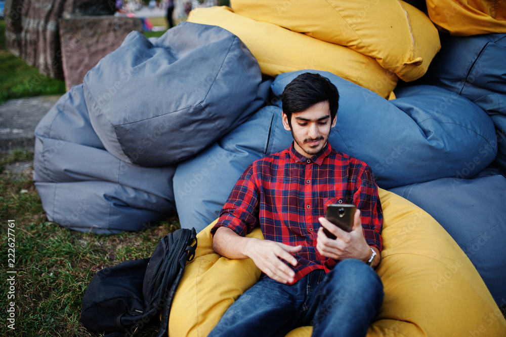 Fototapeta premium Young indian student man at checkered shirt and jeans sitting and relax at outdoor pillows. Spending time with mobile phone.