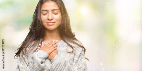 Young beautiful arab woman wearing winter sweater over isolated background smiling with hands on chest with closed eyes and grateful gesture on face. Health concept. photo