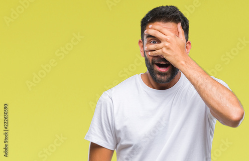 Adult hispanic man over isolated background peeking in shock covering face and eyes with hand, looking through fingers with embarrassed expression.