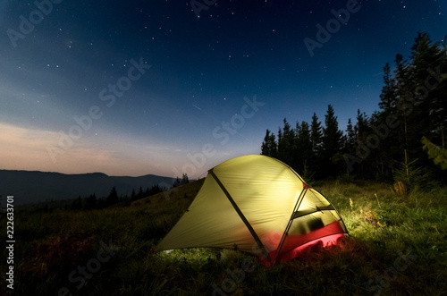 Astrophotography of night camp and starry skies at Carpatian mountains in Ukraine. Green tent on the foreground is highlighted from the inside. Concept of backpacking travel lifestyle.