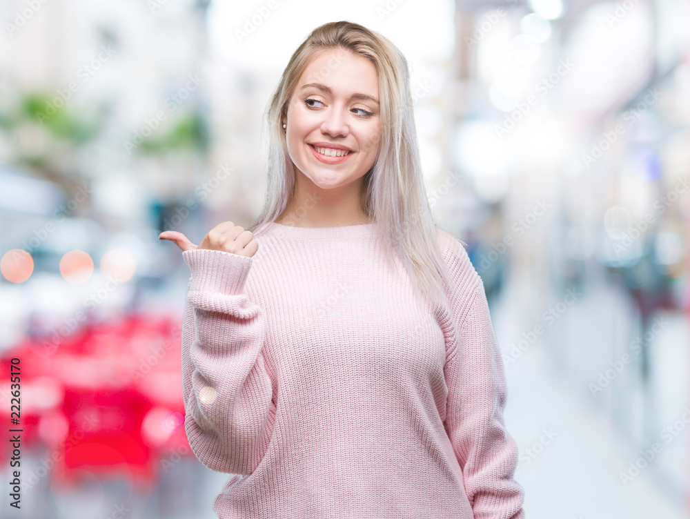 Young blonde woman wearing winter sweater over isolated background smiling with happy face looking and pointing to the side with thumb up. Young blonde woman wearing winter sweater over isolated background smiling with happy face looking and pointing to the side with thumb up.