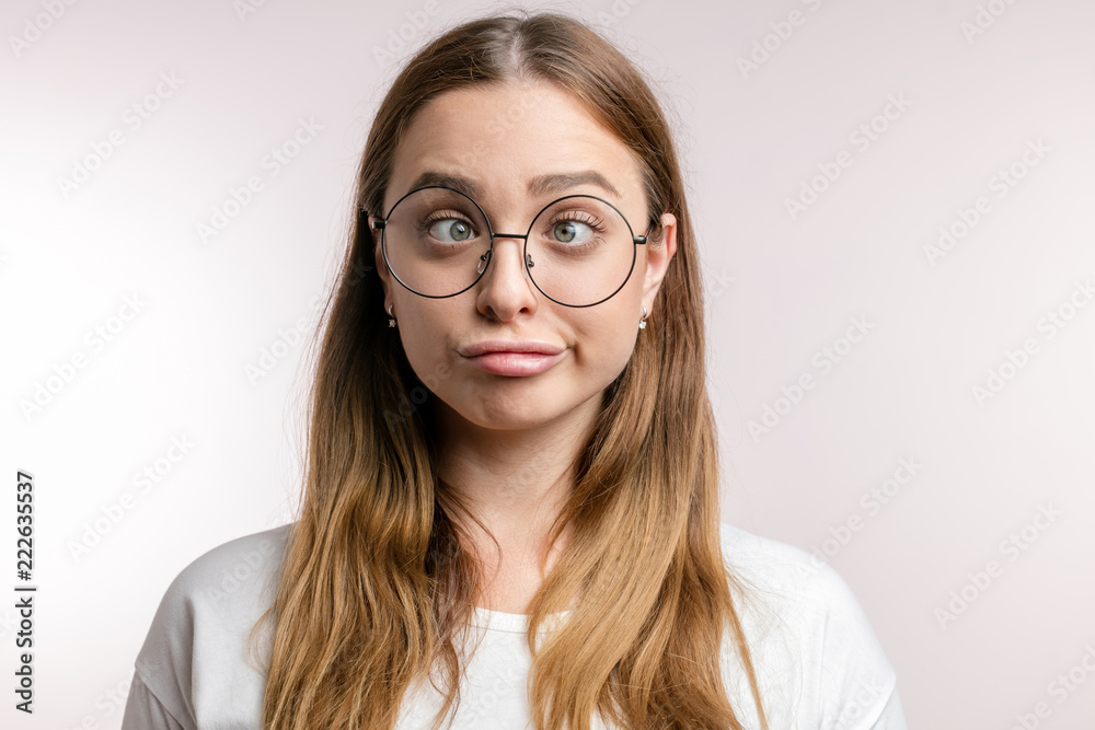Naklejka premium fair-haired attractive girl makes grimace, woman in round glasses crossing eyes student is tired after exams.woman in white T-shirt maks stupid face. closeup portrait