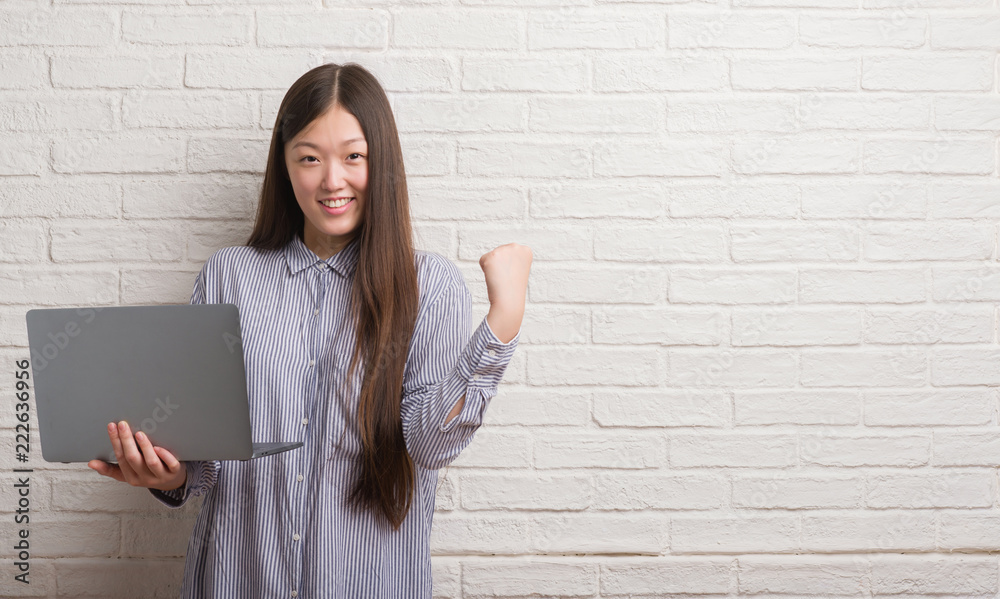 Young Chinese woman over brick wall using laptop screaming proud and celebrating victory and success very excited, cheering emotion Young Chinese woman over brick wall using laptop screaming proud and celebrating victory and success very excited, cheering emotion