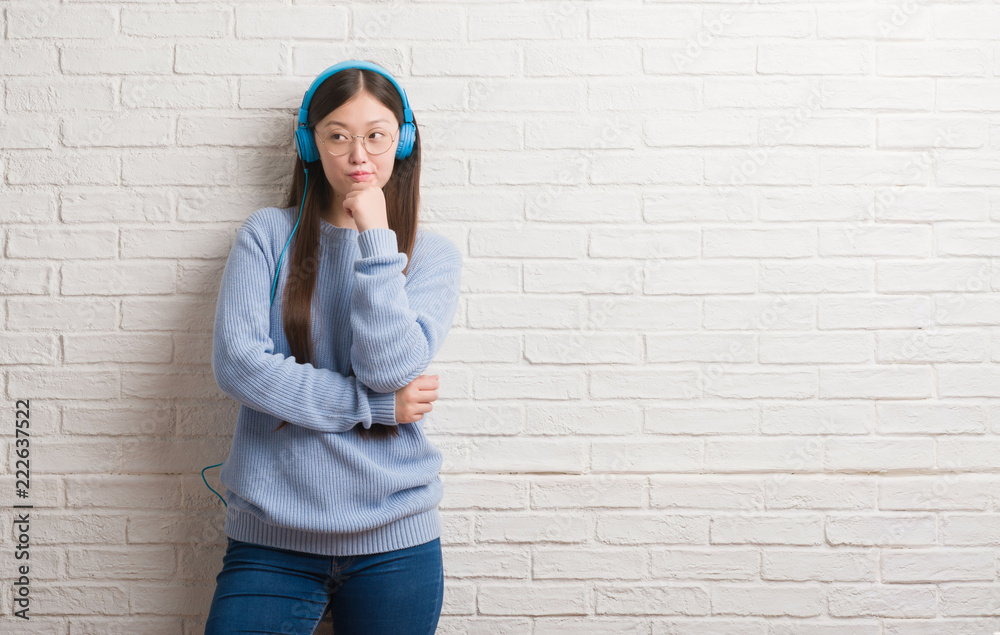 Young Chinese woman over brick wall wearing headphones serious face thinking about question, very confused idea