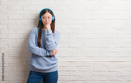 Young Chinese woman over brick wall wearing headphones serious face thinking about question, very confused idea © Krakenimages.com