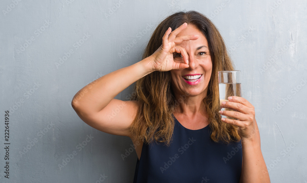 Middle age hispanic woman drinking glass of water with happy face smiling doing ok sign with hand on eye looking through fingers Middle age hispanic woman drinking glass of water with happy face smiling doing ok sign with hand on eye looking through fingers