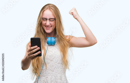 Blonde teenager woman holding smartphone and wearing headphones annoyed and frustrated shouting with anger, crazy and yelling with raised hand, anger concept