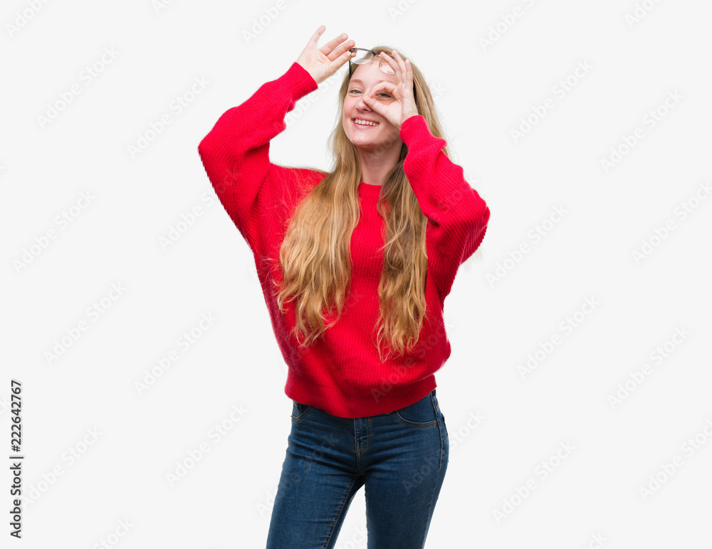 Blonde teenager woman wearing red sweater doing ok gesture with hand smiling, eye looking through fingers with happy face. Blonde teenager woman wearing red sweater doing ok gesture with hand smiling, eye looking through fingers with happy face.