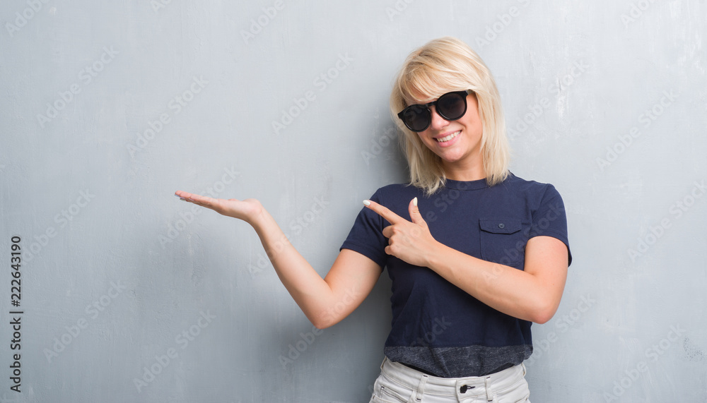 Adult caucasian woman over grunge grey wall wearing sunglasses amazed and smiling to the camera while presenting with hand and pointing with finger. Adult caucasian woman over grunge grey wall wearing sunglasses amazed and smiling to the camera while presenting with hand and pointing with finger.