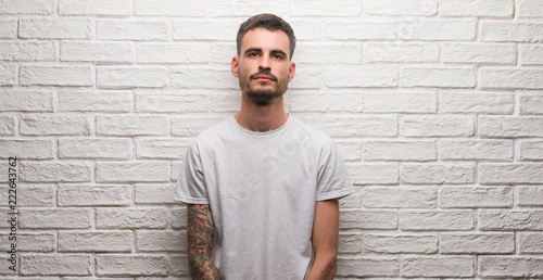 Young adult man standing over white brick wall with a confident expression on smart face thinking serious