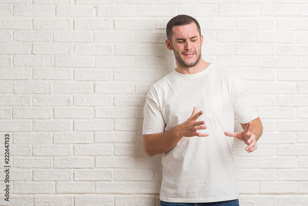 Young caucasian man standing over white brick wall angry and mad raising fist frustrated and furious while shouting with anger. Rage and aggressive concept. Young caucasian man standing over white brick wall angry and mad raising fist frustrated and furious while shouting with anger. Rage and aggressive concept.