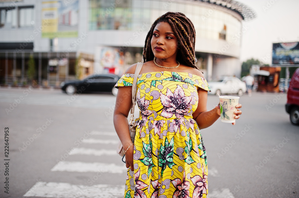 Cute small height african american girl with dreadlocks, wear at coloured yellow dress, walking at crosswalk against trade center with cup of coffee at hand. Cute small height african american girl with dreadlocks, wear at coloured yellow dress, walking at crosswalk against trade center with cup of coffee at hand.