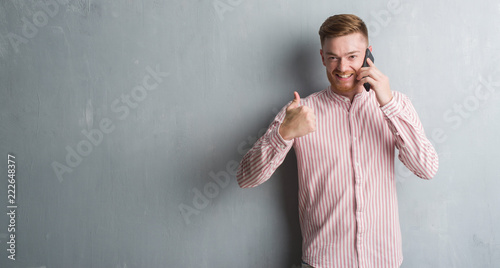 Young redhead man over grey grunge wall talking on the phone happy with big smile doing ok sign, thumb up with fingers, excellent sign