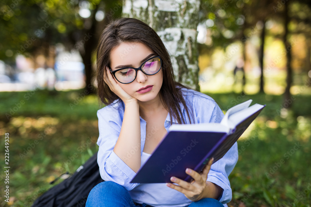 Fototapeta premium Beautiful girl reading a book with bored emotion in summer park