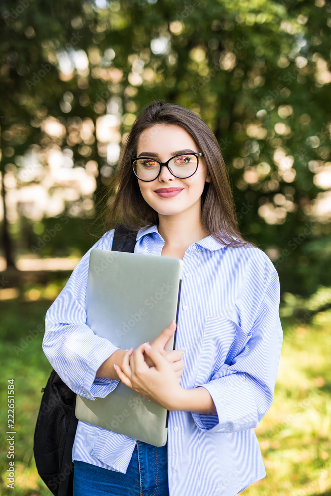 Smiling brunette student wearing glasses and shirt walking outdoor with laptop and backpack background of green park Smiling brunette student wearing glasses and shirt walking outdoor with laptop and backpack background of green park