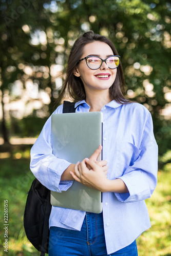 Smiling brunette student wearing glasses and shirt walking outdoor with laptop and backpack background of green park