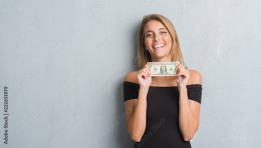 Beautiful young woman over grunge grey wall holding a dollar with a happy face standing and smiling with a confident smile showing teeth