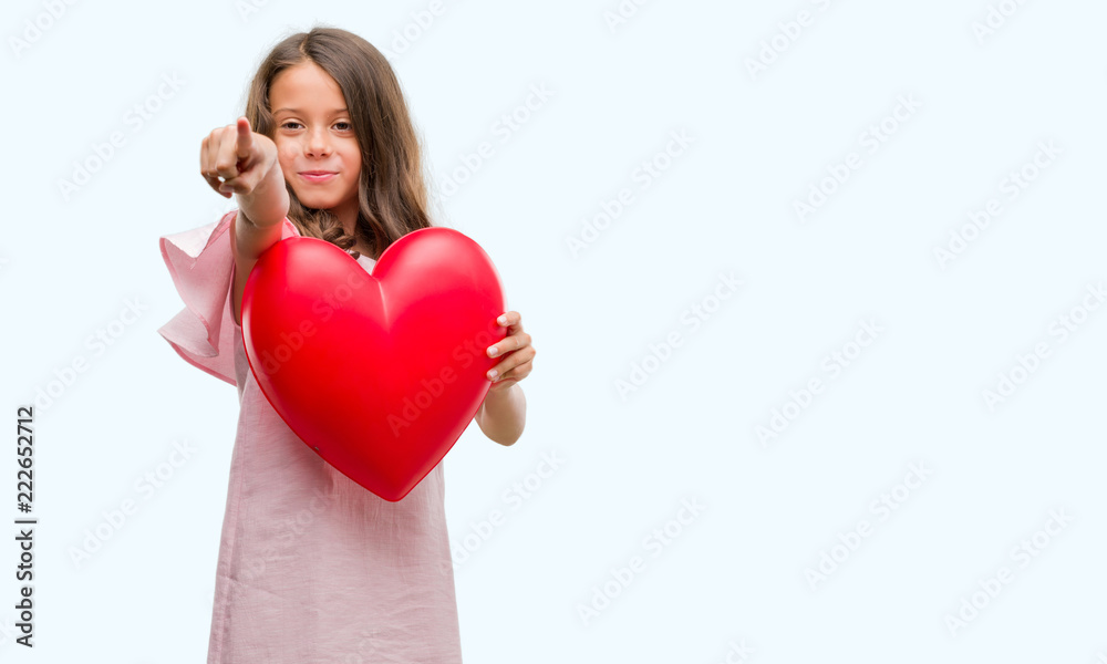 Brunette hispanic girl holding red heart pointing with finger to the camera and to you, hand sign, positive and confident gesture from the front Brunette hispanic girl holding red heart pointing with finger to the camera and to you, hand sign, positive and confident gesture from the front