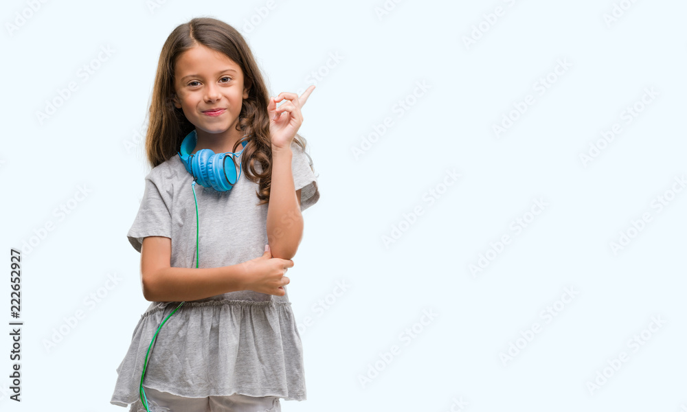 Brunette hispanic girl wearing headphones very happy pointing with hand and finger to the side Brunette hispanic girl wearing headphones very happy pointing with hand and finger to the side