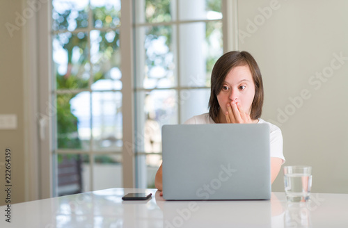 Down syndrome woman at home using computer laptop cover mouth with hand shocked with shame for mistake, expression of fear, scared in silence, secret concept