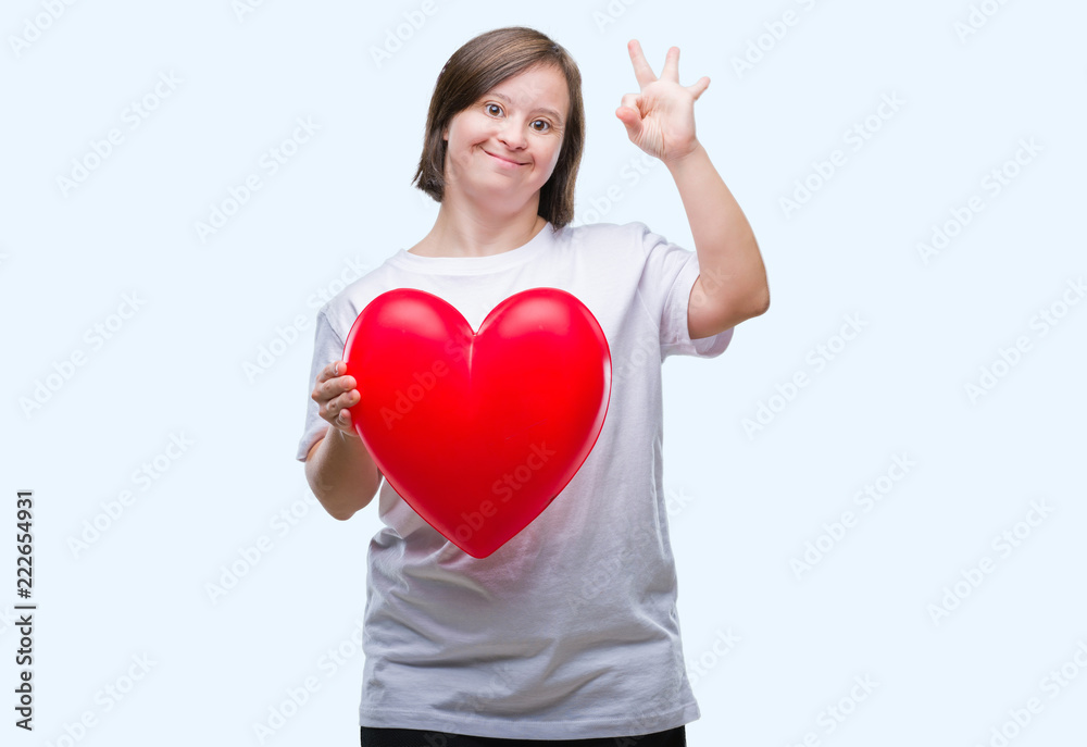 Young adult woman with down syndrome holding red heart over isolated background doing ok sign with fingers, excellent symbol Young adult woman with down syndrome holding red heart over isolated background doing ok sign with fingers, excellent symbol