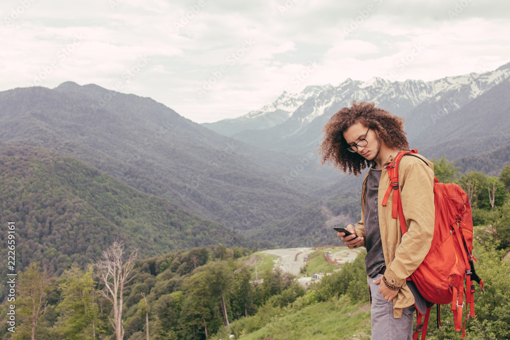 Male hiker with travel backpack search route on mobile navigator while walking near high mountains