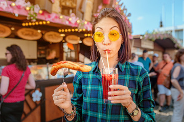 Young happy asian traveler woman eating fried sausage and drinks mug of beer at the fair market square in Germany, beer and food festival concept