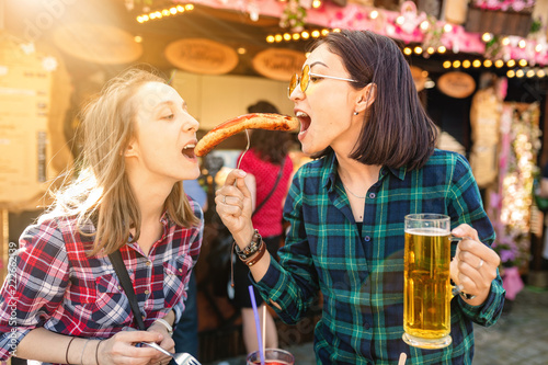 Two girls friends having fun and eating fried sausage and drinks mug of beer at the fair market square in Germany, beer and local food festival concept