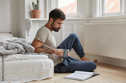 Relaxed concentrated bearded man enjoys reading detective story, focused in book, sits on floor in bedroom, holds mug with coffee or tea, has beard, enriches his knowledge in financial sphere