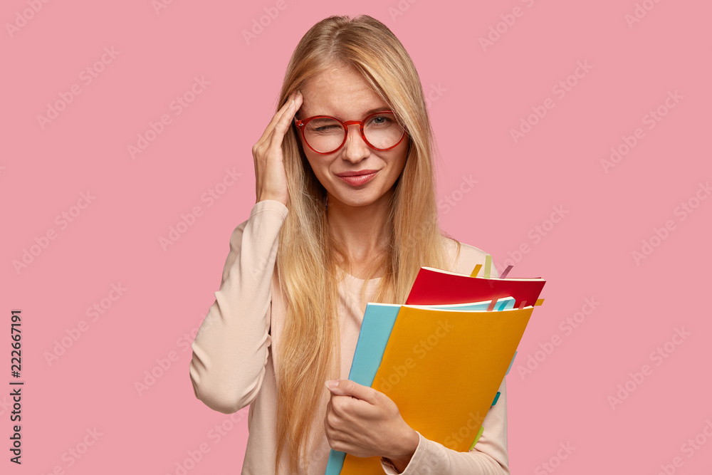 Sad forgetful tired pretty pupil keeps hand on temple, has light long hair, wears glasses, suffers from headache, stands against pink background. Youth, studying, tiredness and lifestyle concept Sad forgetful tired pretty pupil keeps hand on temple, has light long hair, wears glasses, suffers from headache, stands against pink background. Youth, studying, tiredness and lifestyle concept